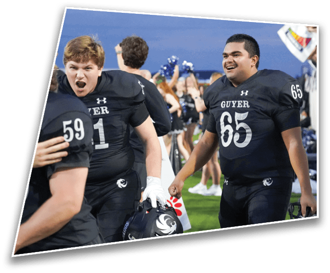 Two people wearing black football uniforms with the word “GUYER" and numbers 65 and 91 are smiling and interacting on a sports field with cheerleaders in the background. Description generated by AI