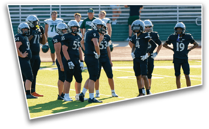 Several people wearing black football uniforms and helmets stand on a green field, appearing to be in a huddle or discussion during a game or practice. Description generated by AI
