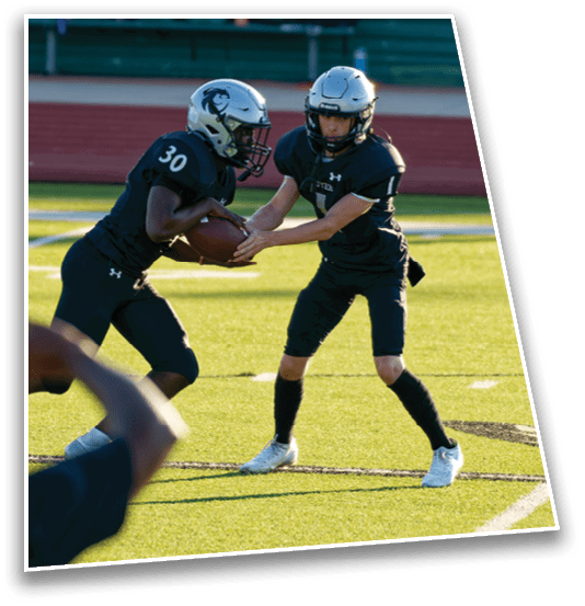Two people in football uniforms and helmets are on a field with one handing a football to the other during a game or practice. Description generated by AI