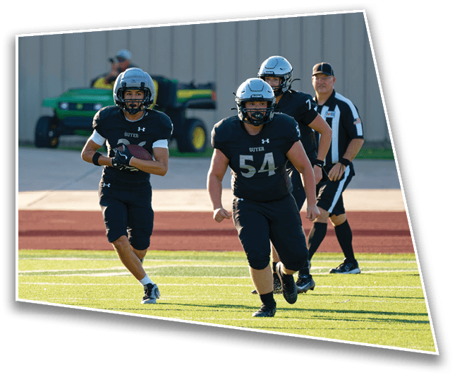 Three people in black football uniforms and helmets are running on a field with a referee walking behind them. Description generated by AI