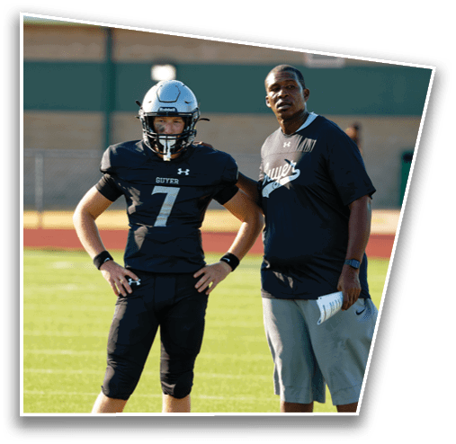 Two people wearing black sports uniforms with “Guyer" on them stand outdoors, one in a football helmet and jersey number seven, and the other holding a football. Description generated by AI