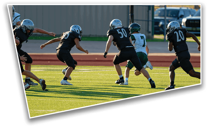 Four people in black sports uniforms are running toward a person in a white and green sports uniform on a grassy field during a game. Description generated by AI