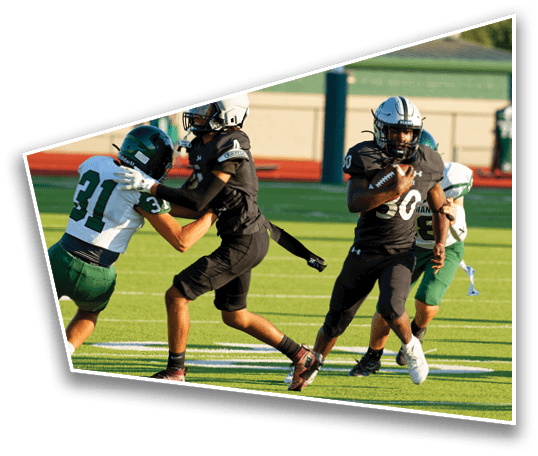 Two people in black sports uniforms are playing football on a field while two others in green and white uniforms attempt to block or tackle them. Description generated by AI