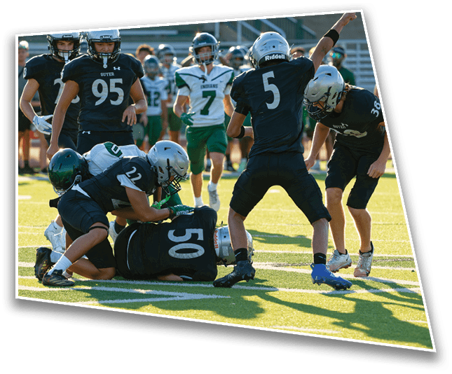 Several people wearing football uniforms are engaged in a play on a sunlit field with one person on the ground and others surrounding them. Description generated by AI
