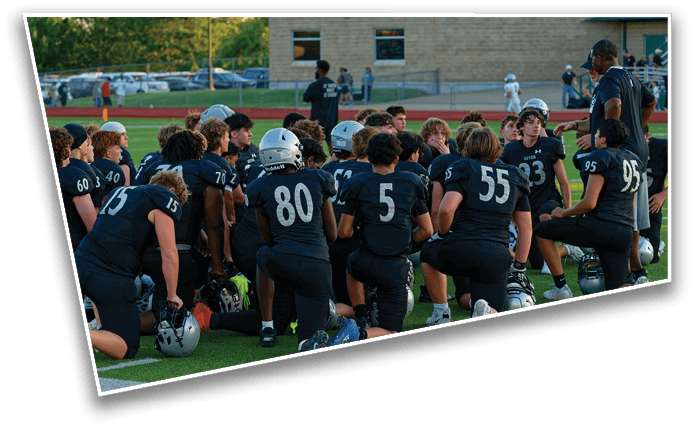 A group of people wearing black football uniforms with helmets gathered around a coach on a sports field during a game or practice. Description generated by AI