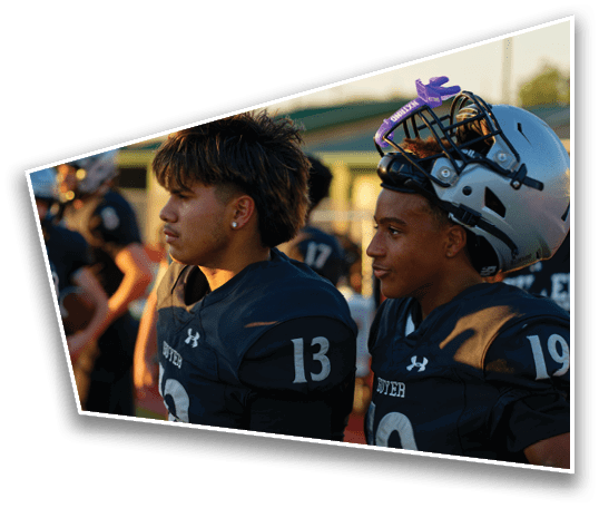 A group of people wearing football helmets and uniforms are gathered outdoors during sunset, with one person in the foreground looking to the side. Description generated by AI