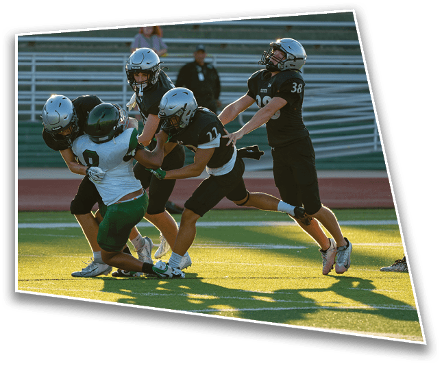 Several people wearing helmets and sports uniforms are actively engaged in a football game on a sunlit field with bleachers in the background. Description generated by AI