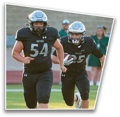 Two people wearing football helmets and black jerseys with the word “GUYER" are actively engaged in a football game, with one holding a football. Description generated by AI