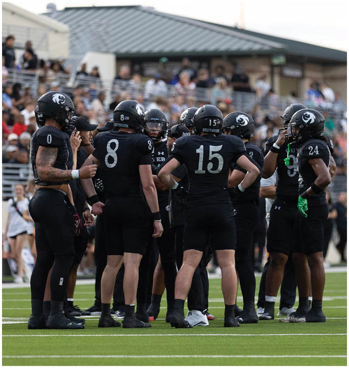 A group of people wearing black football uniforms and helmets are gathered on a field, seemingly discussing strategy during a game. Description generated by AI