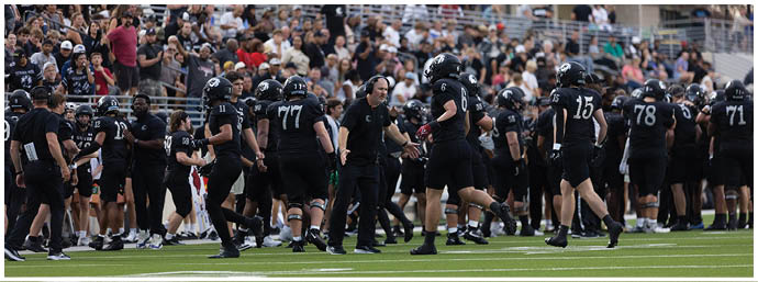 A group of people wearing black sports uniforms and helmets are gathered on a football field with a crowd watching from the stands. Description generated by AI