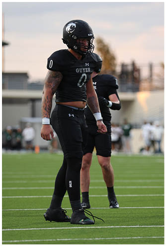 A person wearing a black football uniform and helmet with the word “GUYER" and the number 0 stands on a field during sunset. Description generated by AI