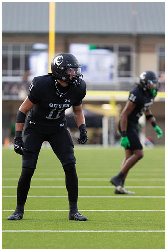 An adult wearing a black football uniform and helmet with the word “GUYER" and the number 11 is in a ready stance on a football field. Description generated by AI
