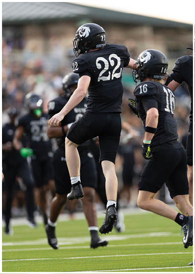 A person wearing a black football uniform with the number 22 is in a dynamic pose on a field with other players in the background. Description generated by AI