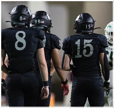 A person wearing a dark football uniform with the number 15 stands facing three people in white football uniforms, one of whom is pointing forward on a sports field. Description generated by AI