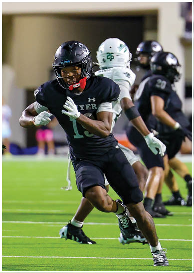 An adult in a black football uniform is running with the ball while being pursued by an adult in a white football uniform on a green field during a game. Description generated by AI