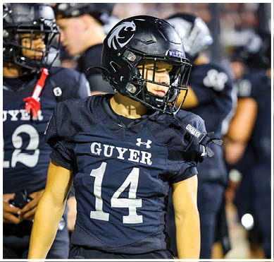 A person wearing a black football uniform with the number 14 and the word “GUYER" on the jersey is standing on a field with other players in the background. Description generated by AI
