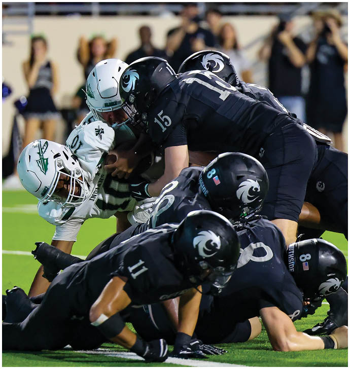 Two people in dark football uniforms are on the ground during a game on a green field. Description generated by AI