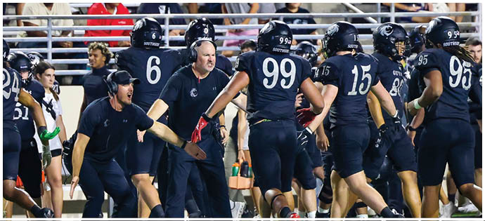A group of people wearing dark sports uniforms and helmets are gathered on a field with some individuals appearing to be coaches giving instructions. Description generated by AI