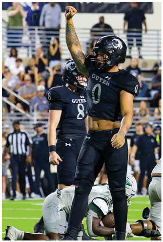 A person wearing a black football uniform and helmet with “Guyer" on the jersey raises a tattooed arm in celebration on a football field. Description generated by AI