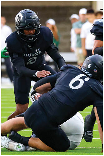 Two football players in dark uniforms are engaged in a tackle on a green field with blurred spectators in the background. Description generated by AI