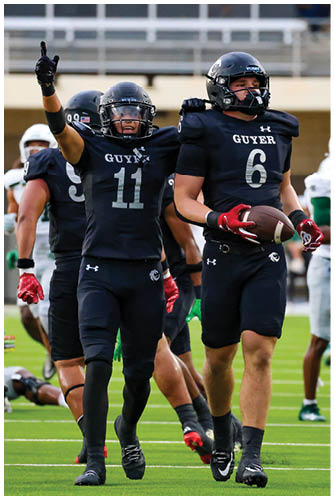A person in a black football uniform holding a football is walking on a field while two people in white football uniforms stand in the background. Description generated by AI