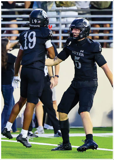 Two people in black football uniforms celebrate on a field near the end zone while others and cheerleaders watch in the background. Description generated by AI