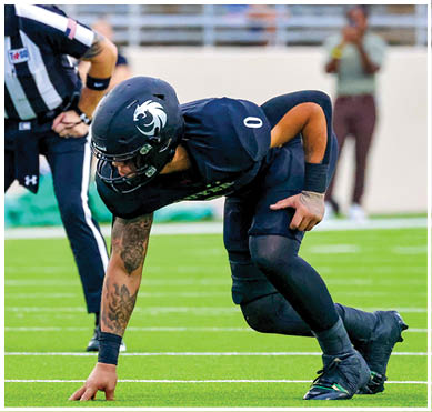 A person in athletic clothing is crouched on a green field with one hand touching the ground and another person stands nearby. Description generated by AI