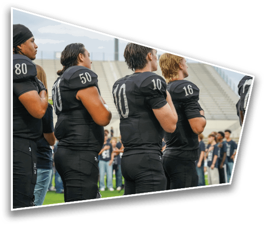 Four people wearing black football uniforms with numbers on their backs stand side by side in a stadium with a crowd in the background. Description generated by AI