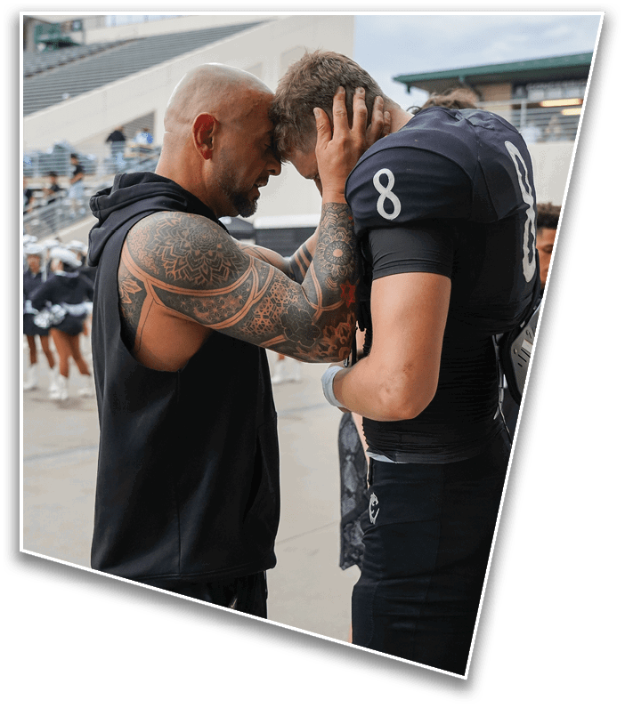An adult with tattoos on their arm is holding the head of a person wearing a black sports uniform with the number 8, appearing to offer comfort or encouragement on a sports field. Description generated by AI