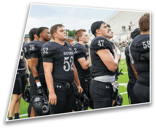 A group of people wearing black football uniforms stand in a line on a field, some holding helmets and one with a hand over their chest. Description generated by AI