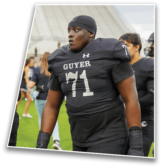 A person wearing a black football uniform with the name “GUYER" and number 71 stands on a field with other players in similar uniforms. Description generated by AI