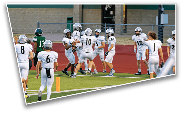 A group of people wearing white football uniforms and helmets are gathered on a sports field near the end zone, appearing to celebrate or strategize during a game. Description generated by AI