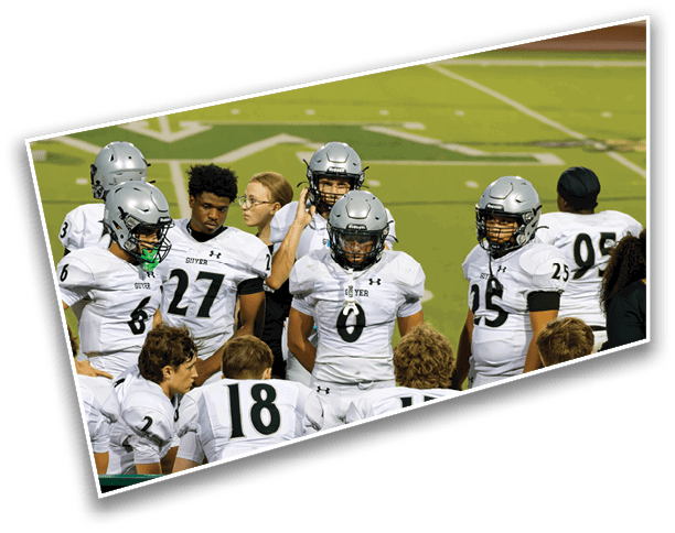 A group of football players in white uniforms and helmets are gathered on a field, some standing and some sitting, while a few adults stand nearby. Description generated by AI