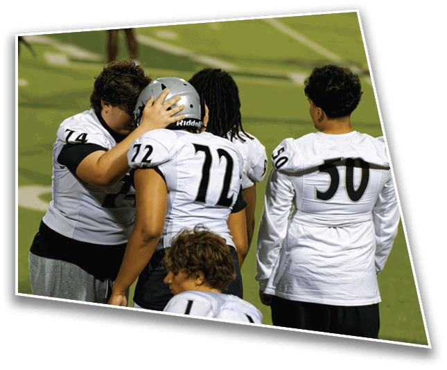 A group of people wearing football uniforms are gathered on a field with one person comforting another by holding their helmeted head. Description generated by AI