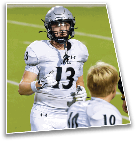 A person wearing a football helmet and white jersey with the number 13 is on a green field, facing another person with short blond hair. Description generated by AI