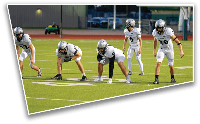 Four people in football uniforms and helmets are positioned on a green field, appearing ready for a play. Description generated by AI