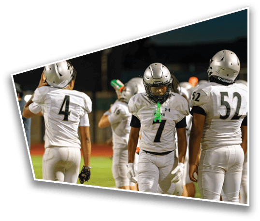 People wearing white football uniforms and helmets are standing on a field at night, with one person in the center wearing number 7 and a green mouthguard hanging from their helmet. Description generated by AI