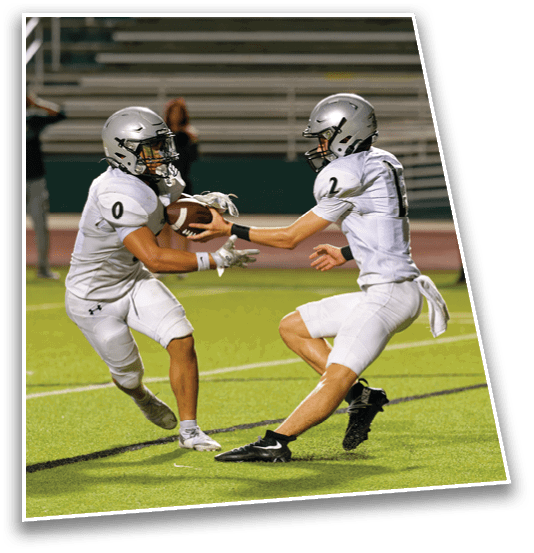 Two people in white football uniforms and helmets are on a field with one handing a football to the other during a game. Description generated by AI