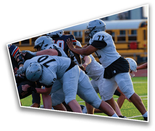 Several people in football uniforms and helmets are engaged in a play on a grassy field with a school bus visible in the background. Description generated by AI