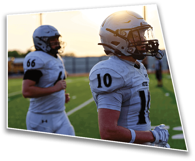 Two people wearing white football uniforms with helmets stand on a green field during sunset. Description generated by AI