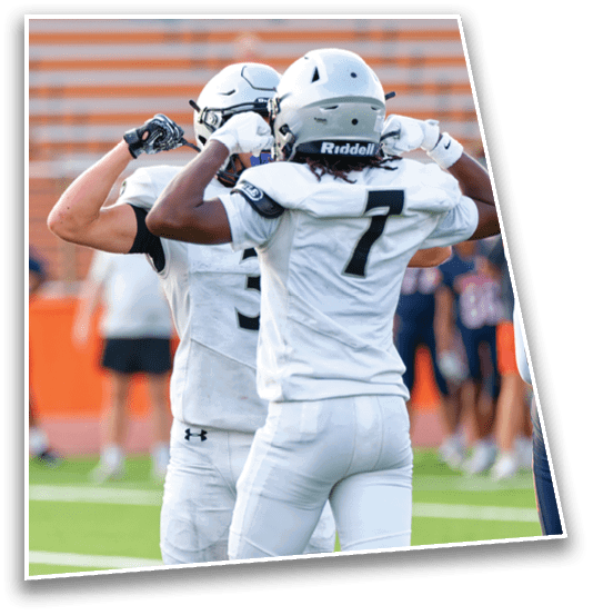 Two people in white football uniforms and helmets are flexing their arm muscles on a football field with blurred spectators in the background. Description generated by AI