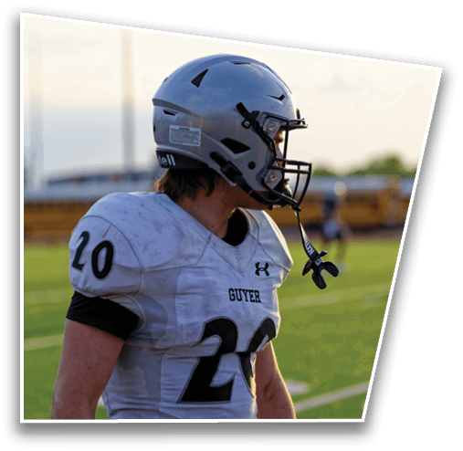 A person wearing a white football uniform with the number 20 and a gray helmet stands on a green field with a blurred background of a school bus and buildings. Description generated by AI