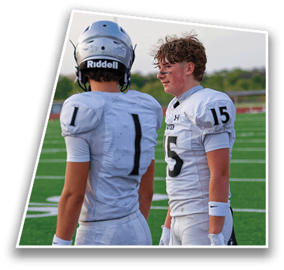 Two people in football uniforms are on a field, one wearing a helmet with the brand name Riddell visible and the other with face paint and curly hair. Description generated by AI