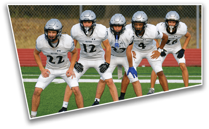 Five people in white football uniforms and helmets are crouched in a ready position on a green field with a red running track and a chain-link fence in the background. Description generated by AI