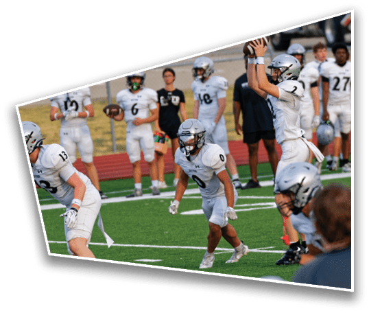 People in white football uniforms are on a green field with one person preparing to throw a football while others are in ready positions. Description generated by AI