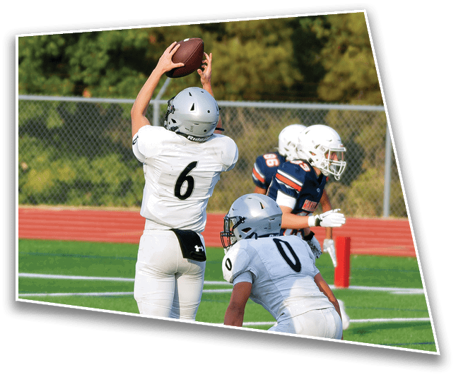 A person wearing a silver helmet and white jersey is catching a football while two other people in white helmets and dark jerseys stand nearby on a field with a chain-link fence and trees in the background. Description generated by AI