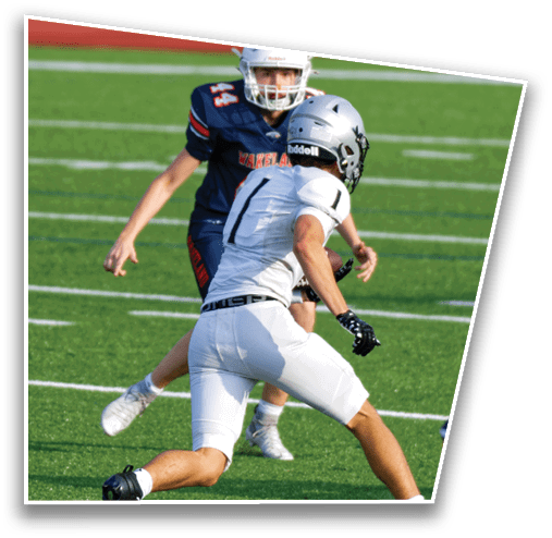Two people in football uniforms are on a field with one person in white crouching and the other in dark blue standing behind. Description generated by AI