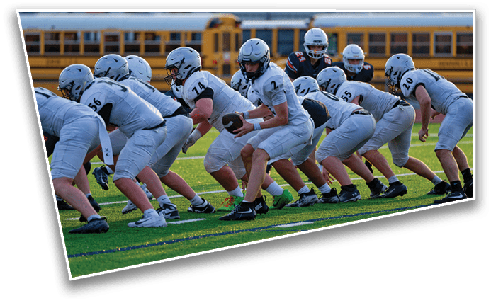 A group of people in white football uniforms are lined up on a field preparing for a play with school buses visible in the background. Description generated by AI