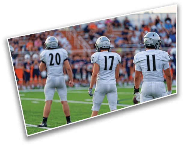 Three people in white football uniforms with helmets stand on a green field facing away from the camera with a blurred crowd in the background. Description generated by AI