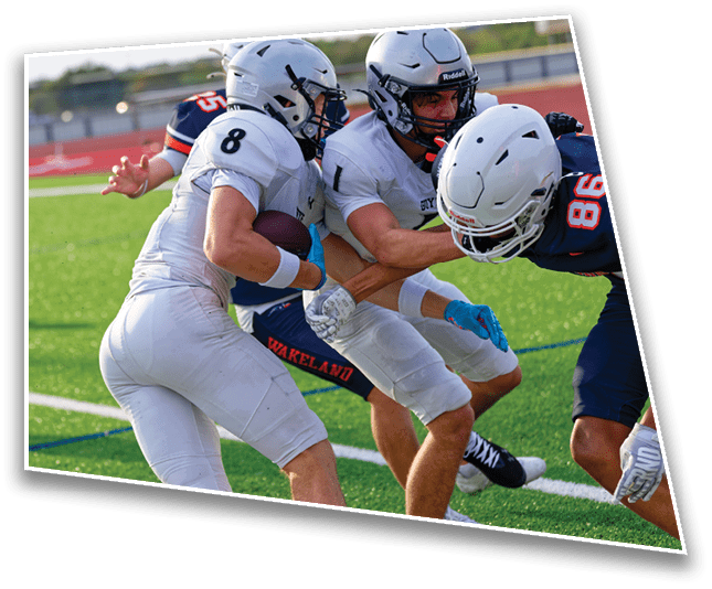 Two people in white football uniforms are running with the ball while being tackled by opponents in dark uniforms on a green field. Description generated by AI